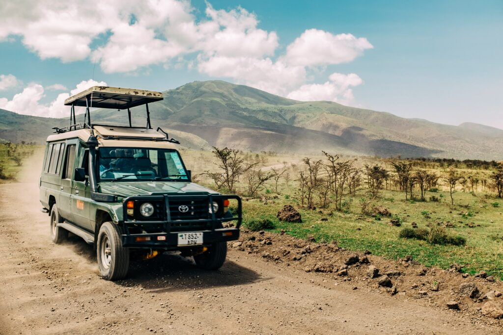 A picture of a sunny day in Africa with a safari truck driving through the park stirring up dust as it drives by. The shrubs on the horizon are short, and there is a green mountain in the back ground with some clouds scattered in the blue sky. The safari truck has a extended roof on top for an added view point.